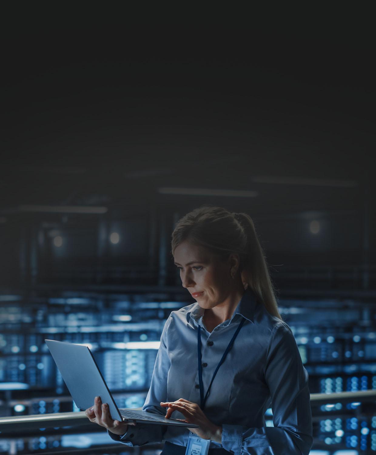 A woman in business causal clothing standing, holding a laptop, in an IT setting.