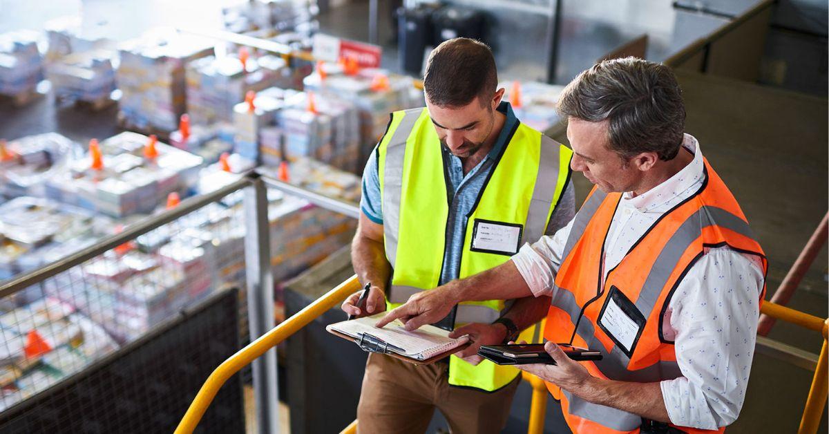 Two men in a warehouse setting wear safety vests and consult a clipboard together.