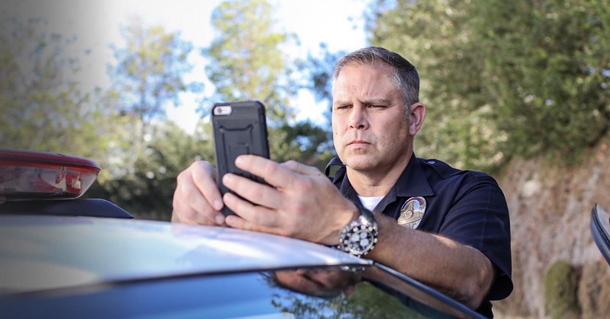 An image of a uniform police officer leaning on his patrol car, staring in concentration at his mobile phone.