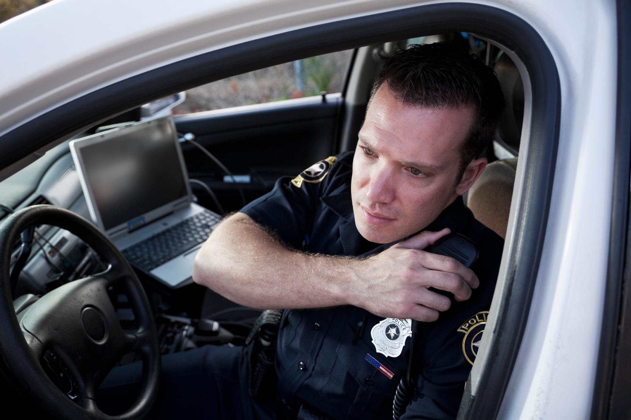 A police officer sitting in car with laptop visible behind him - the officer is speaking into the radio on his shoulder while looking out of the car window.