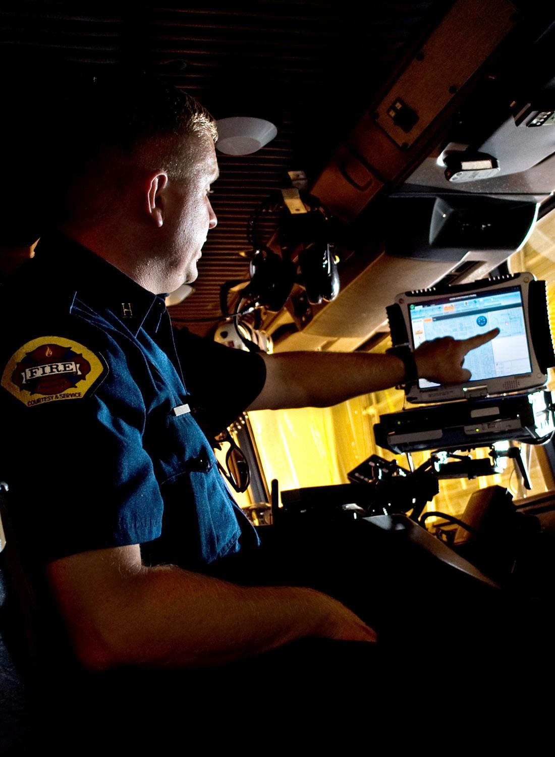 A fire rescue officer in the cab of his fire truck pointing to something on a laptop monitor.