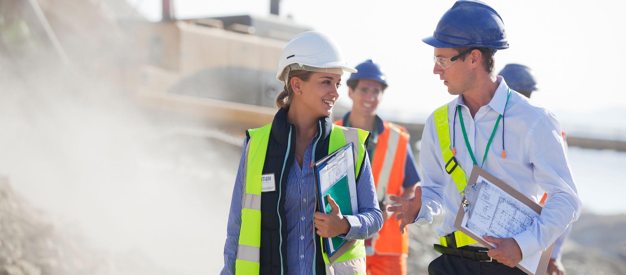 A group of public admin workers appear to be on a construction site. They are wearing hard hats and safety vests, holding clipboards with planning documents smiling and talking.