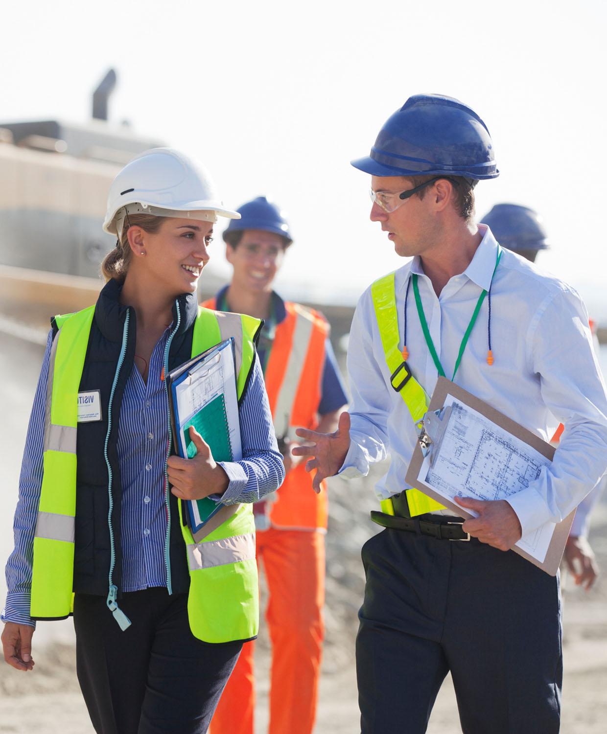 A group of public admin workers appear to be on a construction site. They are wearing hard hats and safety vests, holding clipboards with planning documents smiling and talking.